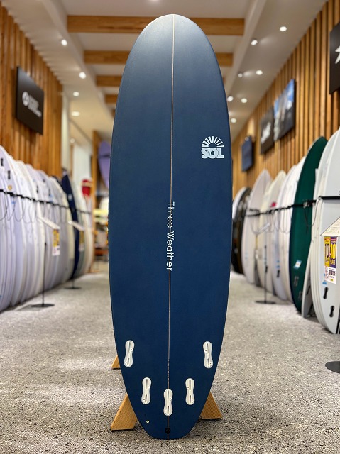 Blue surfboard on a rack in a surf shop, with a SOL logo and vertical 'Free Weather' text near the center.