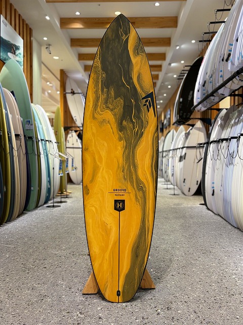a-image4 - SHOP NEWS | ムラサキスポーツオンラインストア 通販 Yellow and black marbled surfboard on a stand in a surf shop aisle, surrounded by racks of other boards.