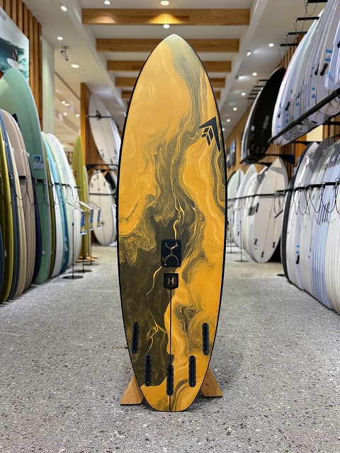 a-image5 - SHOP NEWS | ムラサキスポーツオンラインストア 通販 Yellow and black marbled surfboard standing upright on a wooden display rack in a surf shop with other boards in the background.
