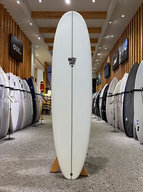 Long white surfboard standing upright on a wooden stand in a surf shop, with racks of boards lining both sides of the aisle.