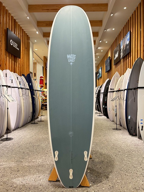 Tall blue-green surfboard on a wooden stand in a showroom, with numerous boards lining racks along the walls.