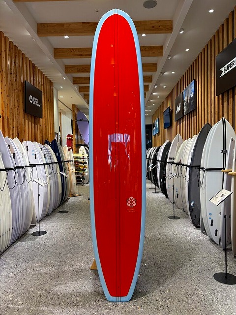 Red surfboard with light blue edging stands upright in a surf shop, with rows of boards lining the walls.