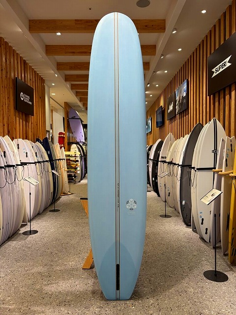 Tall light-blue surfboard standing upright on a display rack inside a surf shop, surrounded by many other boards along wooden walls.