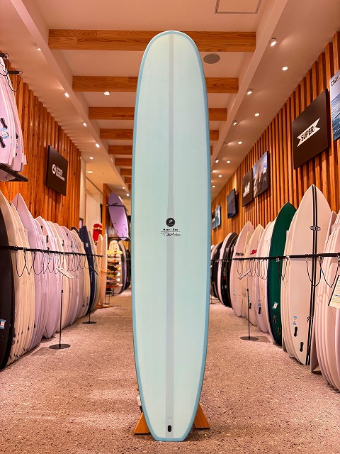a-メイシーブー 1 - SHOP NEWS | ムラサキスポーツオンラインストア 通販 Tall, light blue longboard standing upright on a rack in a surf shop, with rows of surfboards along wooden walls on both sides.