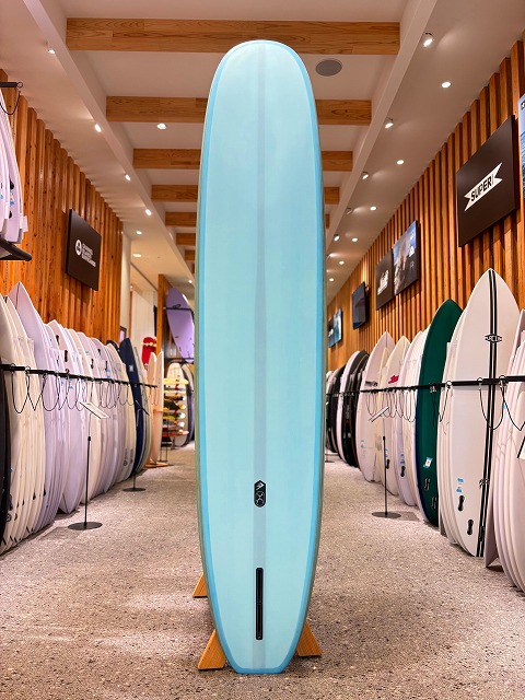 a-メイシーブー 2 - SHOP NEWS | ムラサキスポーツオンラインストア 通販 Tall light-blue surfboard on a wooden stand in a surf shop, with rows of boards lining both walls behind it