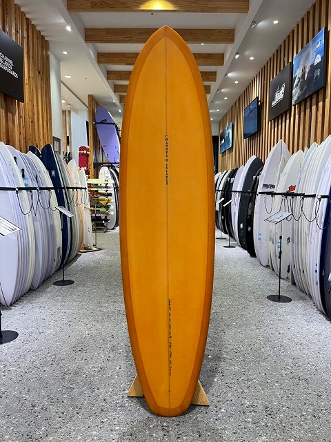Orange surfboard upright on a wooden stand in a surf shop, surrounded by rows of white and colored boards along both walls.