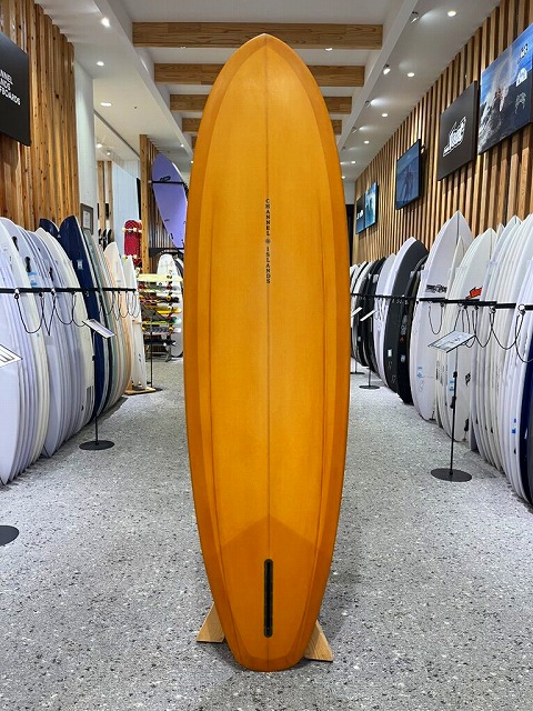 Tall yellow surfboard standing upright on a display rack inside a surf shop, with rows of boards lining the walls and bright lighting above.