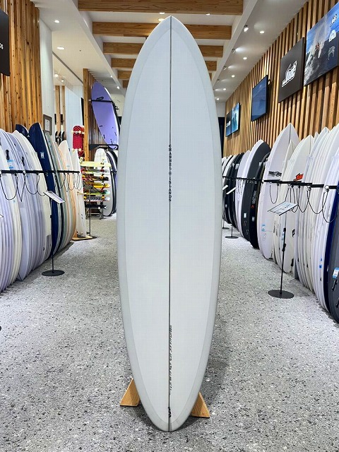 White surfboard standing upright on a wooden stand in a showroom, surrounded by racks of other boards.