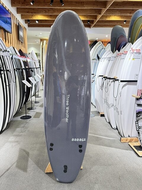 s-IMG_3316 - SHOP NEWS | ムラサキスポーツオンラインストア 通販 Tall gray surfboard standing upright on a stand in a surf shop, surrounded by racks of white and colored boards.