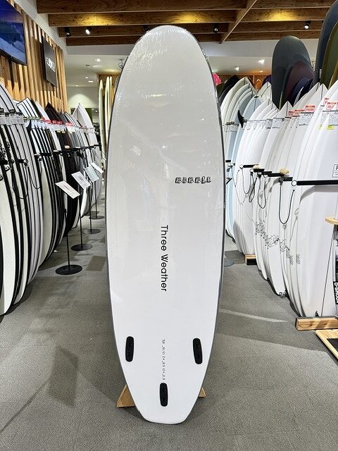 s-IMG_3317 - SHOP NEWS | ムラサキスポーツオンラインストア 通販 White surfboard with three fin boxes standing upright on a display rack in a surf shop, surrounded by numerous other boards; brand name 'Three Weather' visible along the center.
