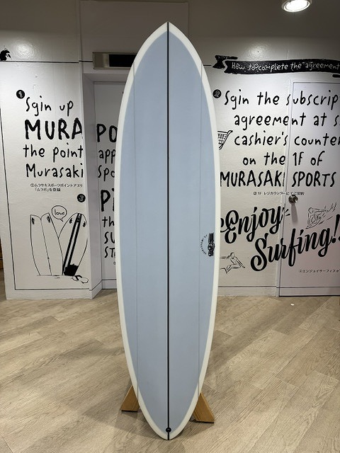 s-IMG_4450 - SHOP NEWS | ムラサキスポーツオンラインストア 通販 Light gray-blue surfboard with a dark center seam, standing upright on a wooden display rack inside a store, surrounded by graffiti-style wall art and signs about surfing.