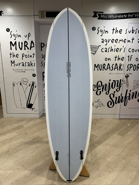 s-IMG_4452 - SHOP NEWS | ムラサキスポーツオンラインストア 通販 Vertical blue-tinted surfboard on a wooden stand in a showroom, with wall graphics in the background