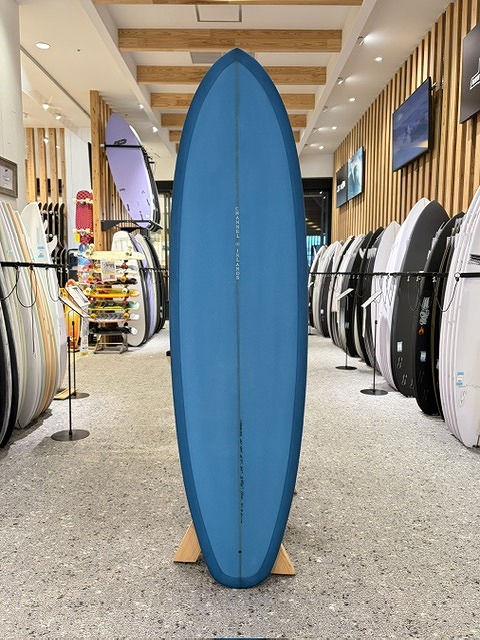 Blue surfboard standing upright on a wooden rack in a surf shop, with other boards visible on both sides inside the showroom.