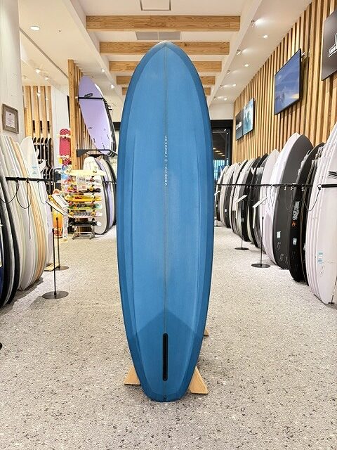 Blue surfboard standing upright on a wooden stand in a surf shop, surrounded by other boards on racks.