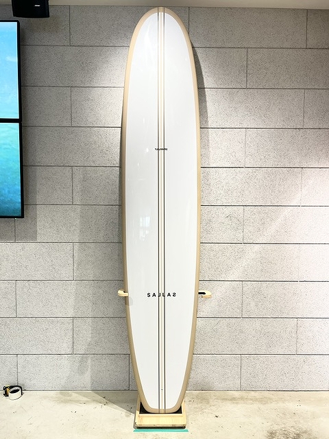 Tall white surfboard with a light wood edge, standing upright on a wooden rack against a gray brick wall indoors.