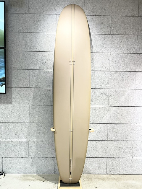 Beige surfboard standing upright on a rack against a gray tiled wall, shown in a showroom or gallery setting.