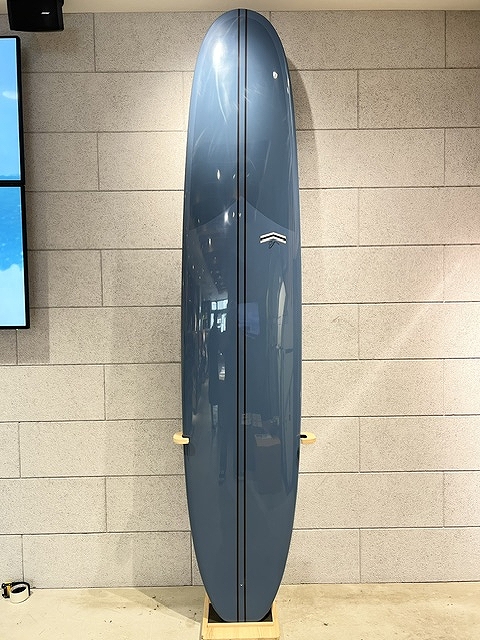 s-IMG_6373 - SHOP NEWS | ムラサキスポーツオンラインストア 通販 Blue-gray surfboard on a wooden stand standing upright against a beige brick wall, featuring a small white chevron logo on the upper half.