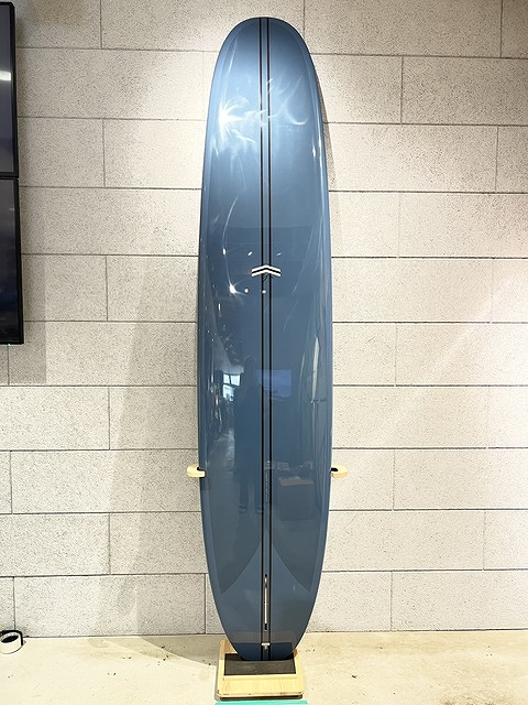 s-IMG_6374 - SHOP NEWS | ムラサキスポーツオンラインストア 通販 Blue surfboard standing upright on a wooden rack against a light gray brick wall, with a vertical center seam and a small logo near the top.