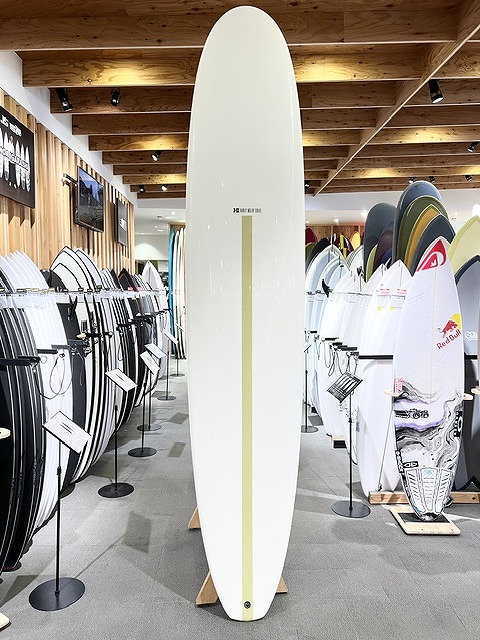 Tall white surfboard on display in a shop, centered among racks of surfboards.