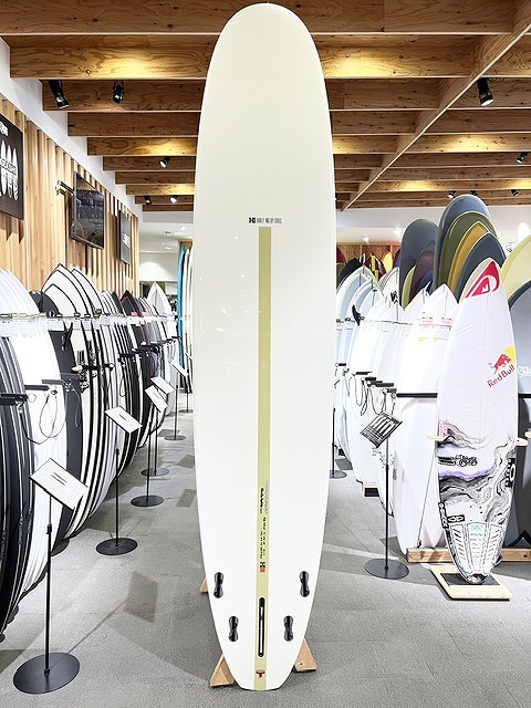 Tall white surfboard standing upright on display in a surf shop, with rows of boards lining the walls and warm wooden beams overhead.