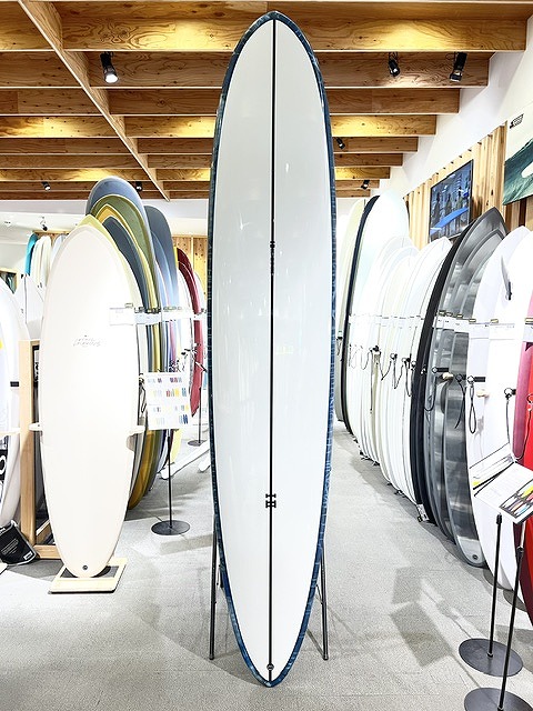Tall white surfboard on a stand in a showroom, surrounded by other boards on racks in a surf shop