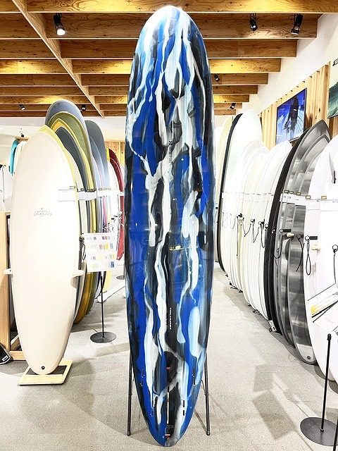 Tall surfboard with blue, white, and black abstract pattern standing upright among other surfboards in a showroom with a wooden-beam ceiling.