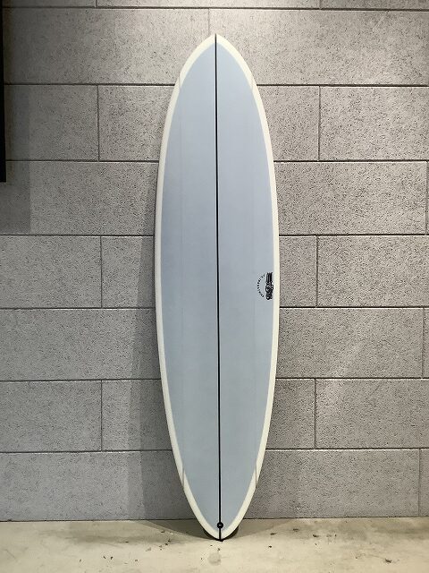 White surfboard with a dark center seam, standing upright against a gray brick wall; small logo near the middle.