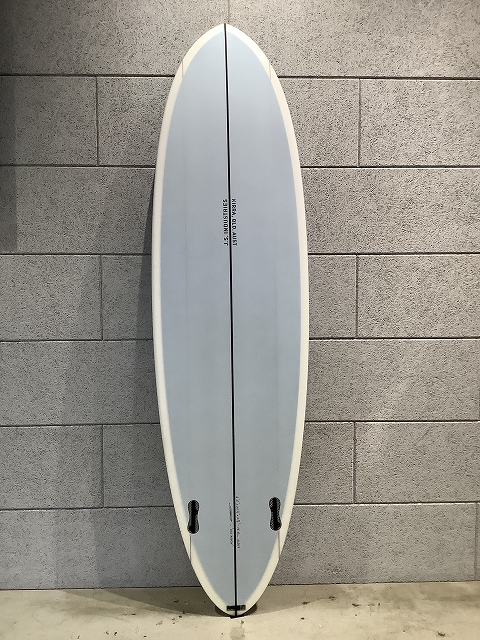 Light blue surfboard with white rails, standing upright against a gray brick wall; a center stringer runs lengthwise with two fin boxes near the bottom.