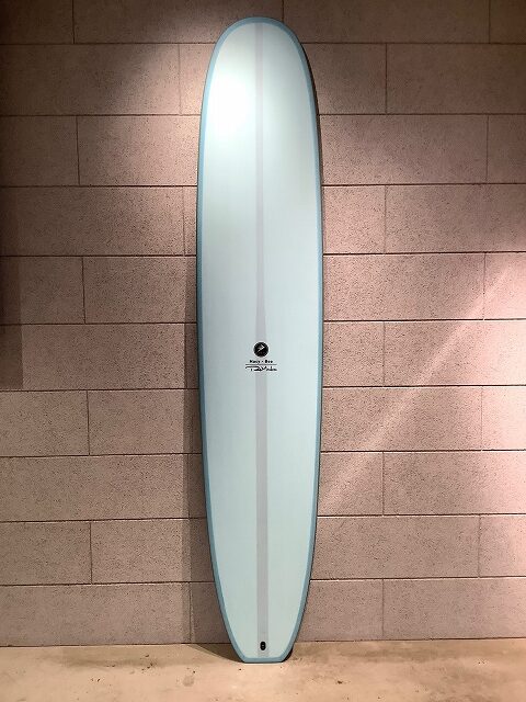 Light blue surfboard standing upright against a tan brick wall in an indoor setting.