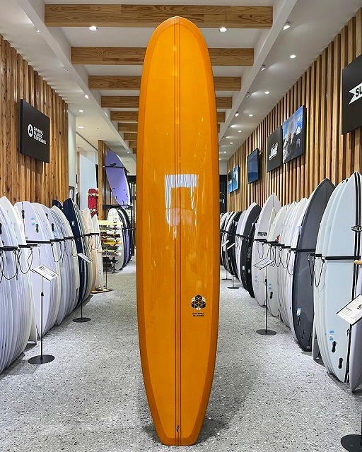 Bright orange longboard standing upright in the center of a surf shop aisle, surrounded by white and colored boards on racks.