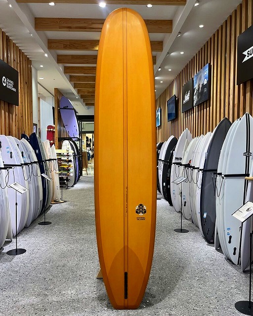 Tall orange surfboard standing upright in a surf shop showroom, surrounded by racks of other surfboards.