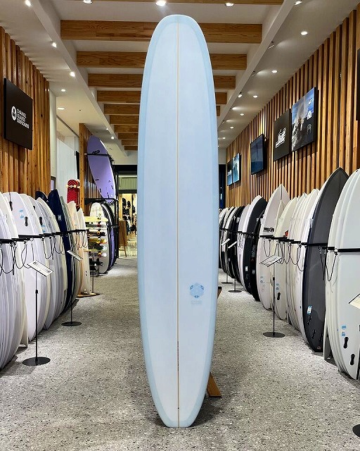 Tall pale blue surfboard standing upright between rows of racks in a surf shop showroom.
