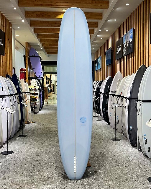 Tall light-blue surfboard standing upright in a surf shop showroom, surrounded by racks of boards.