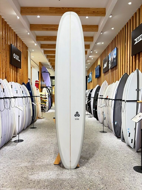 Tall white surfboard standing upright on a display rack in a surf shop showroom, with rows of boards lining both sides and wooden ceiling beams above.