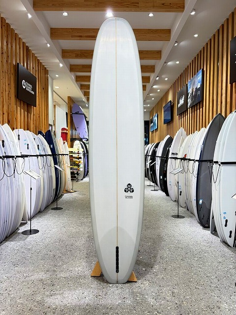 Tall white surfboard standing upright on a wooden stand in a surf shop aisle, with racks of boards on both sides.