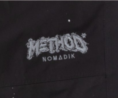 White embroidered logo on dark fabric reading 'METHODS NOMMADIK' (close-up on clothing)