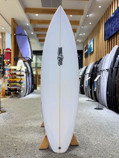 White surfboard on a wooden stand in a surf shop, logo mid-board with racks of boards visible in the background on both sides.