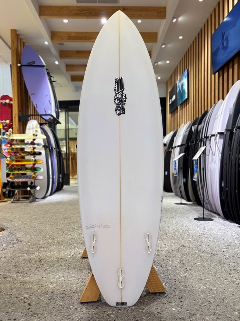White surfboard standing upright on a wooden display rack inside a surf shop, with other boards lining the walls.