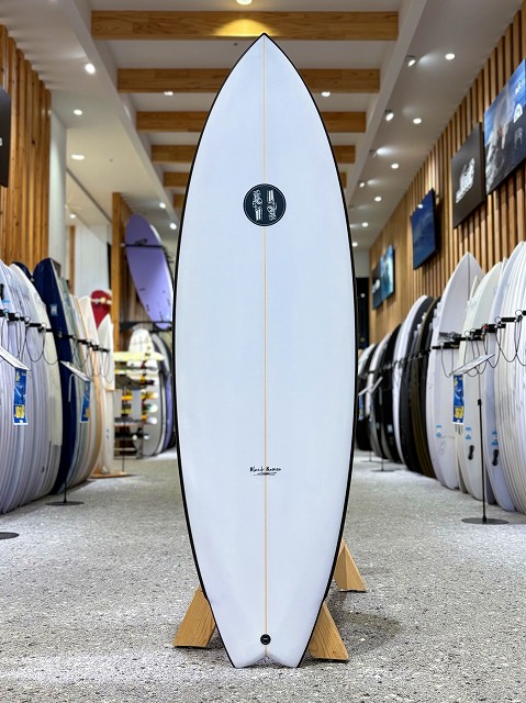 ブラックバロン 1 - SHOP NEWS | ムラサキスポーツオンラインストア 通販 White surfboard with brown edge standing upright on wooden racks in a surf shop, surrounded by other boards on display along the walls.