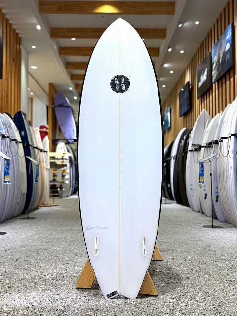 ブラックバロン 2 - SHOP NEWS | ムラサキスポーツオンラインストア 通販 White surfboard on a wooden stand in a surf shop, with a black circular logo near the top and other boards on display nearby.