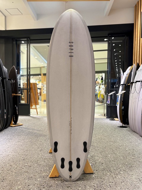 Light-colored surfboard standing upright on display racks in a surf shop, with multiple fin boxes visible along the bottom and other boards in the background.