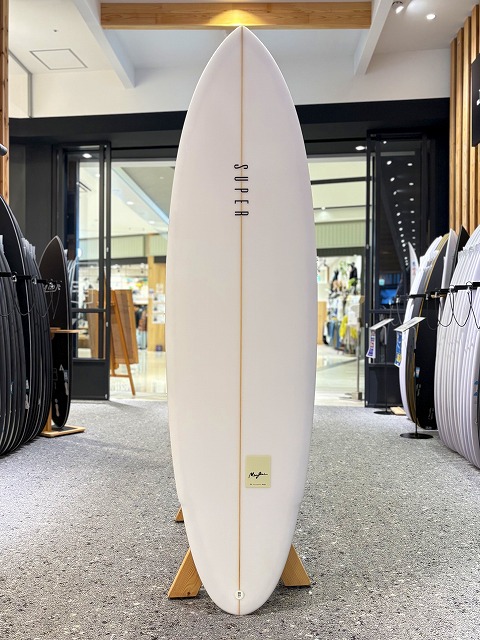 White surfboard on a wooden stand centered in a surf shop, with other boards lined up along the sides.