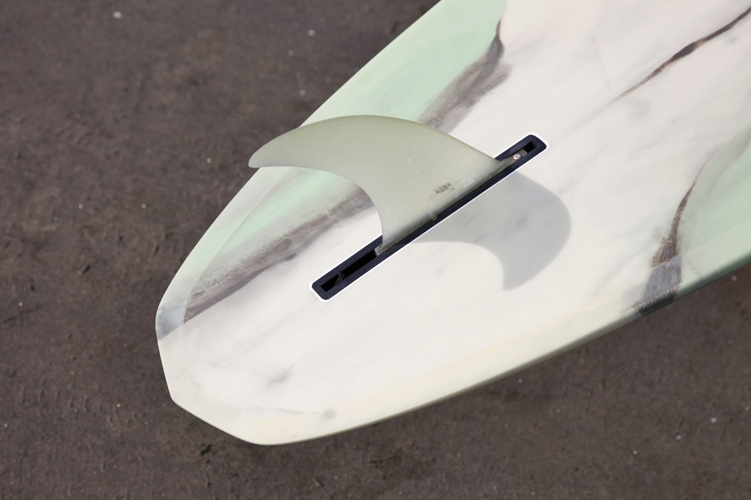 Close-up of a light-colored surfboard tail with a translucent fin in the slot on a dark surface.