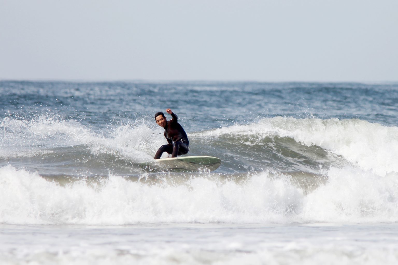 Surfer wearing a black wetsuit rides a wave on a white surfboard near the shore, spray flying around him.