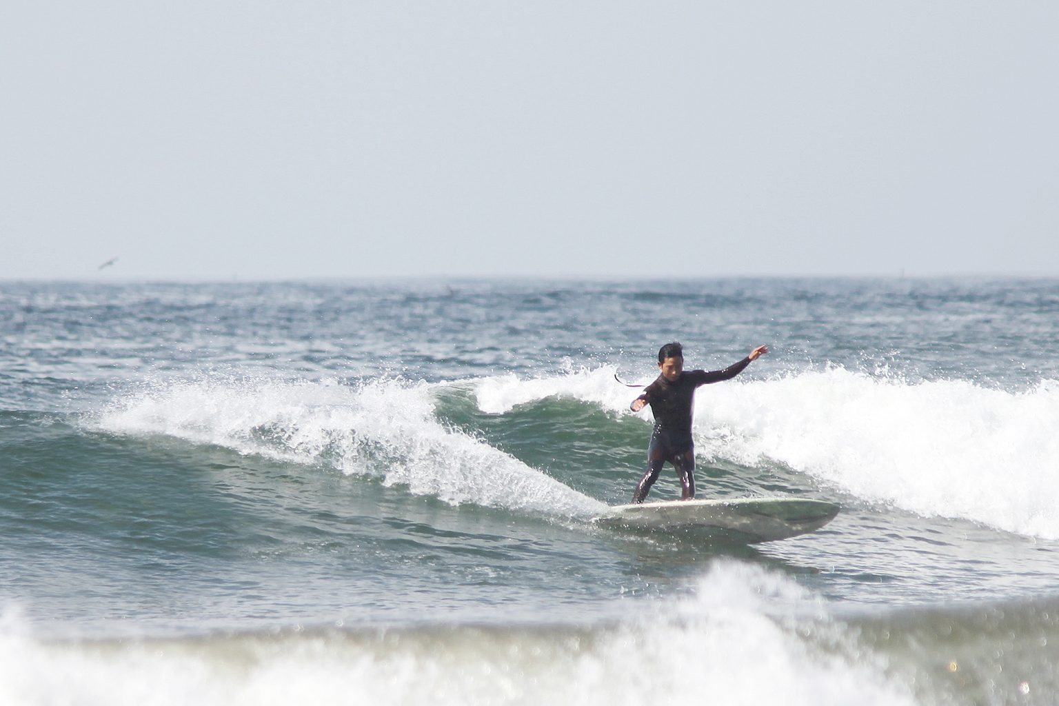 Surfer standing on a board riding a small wave near the shore, wearing a black wetsuit and extending one arm for balance.
