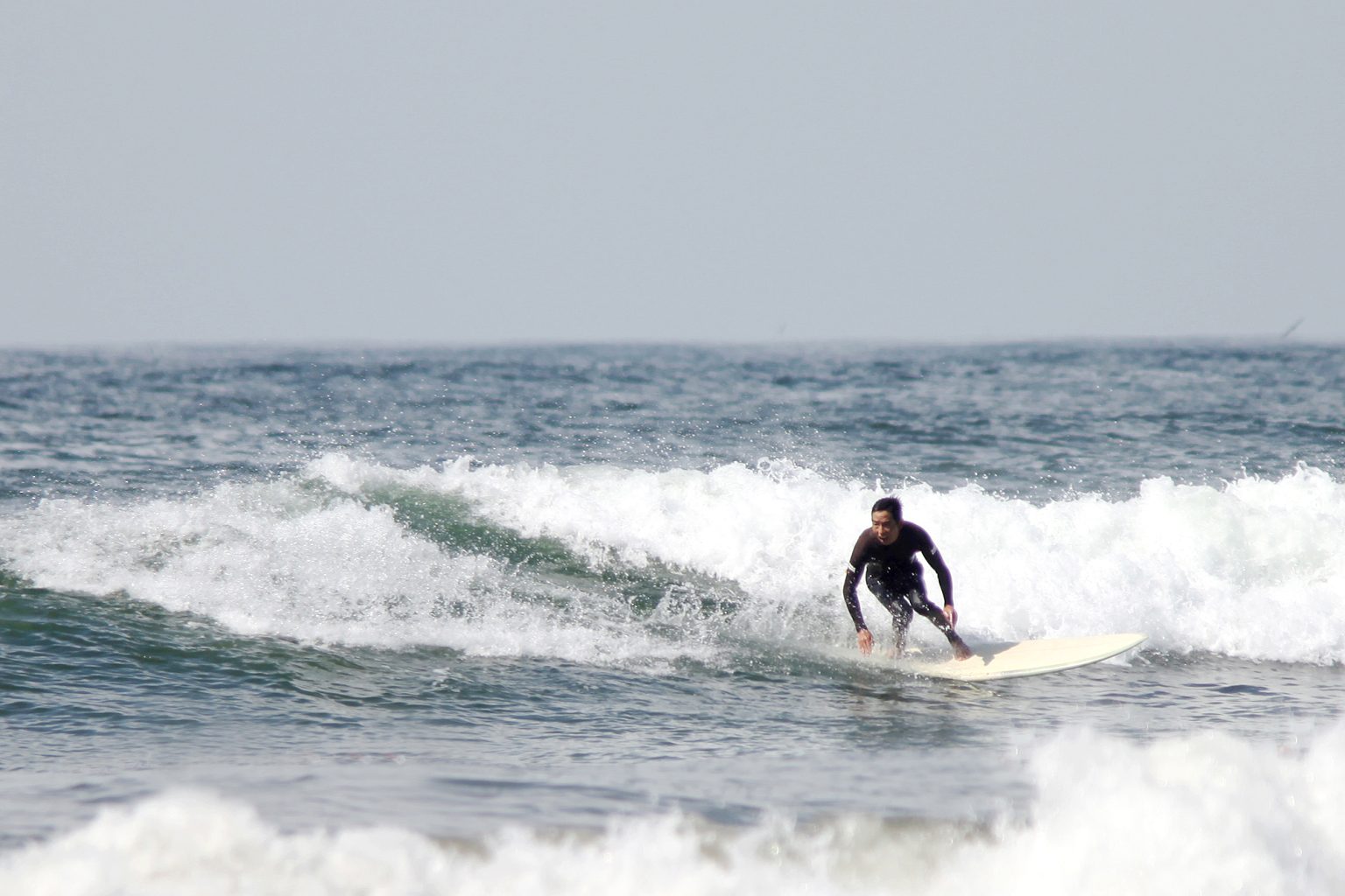 Surfer in a black wetsuit riding a light-colored board on a small breaking wave near shore.