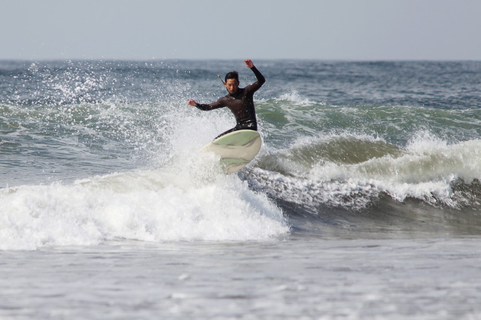 Surfer in a black wetsuit rides a breaking wave with a white-green board, spray flying around the surfer.