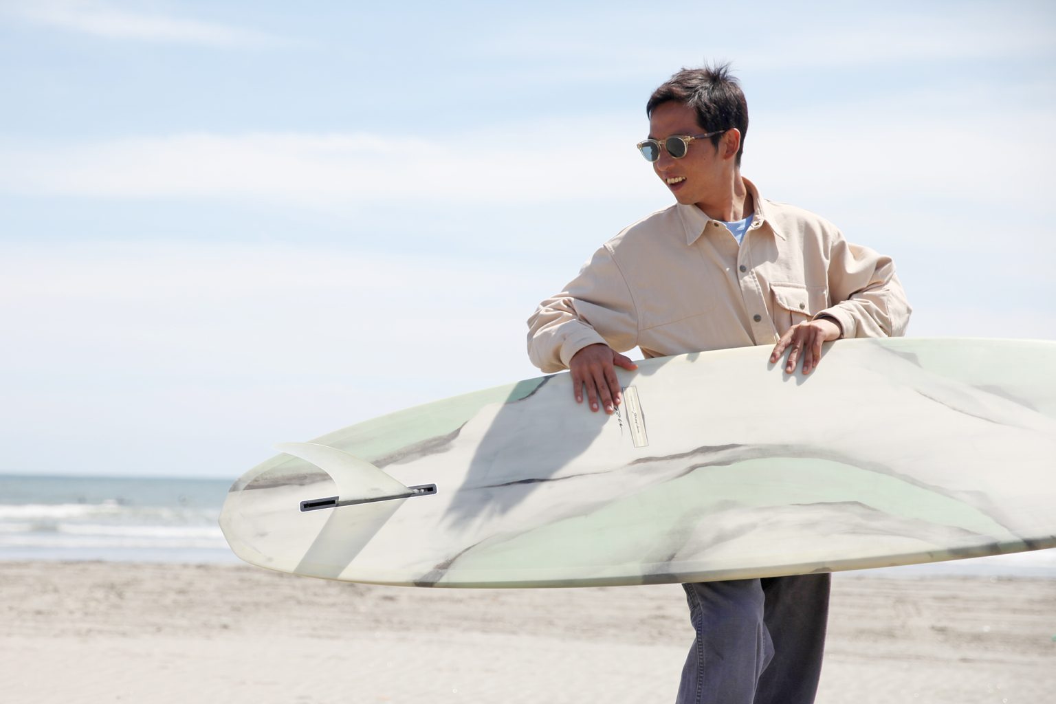 Man at the beach wearing sunglasses and a beige shirt, carrying a surfboard under his arm with the ocean in the background.