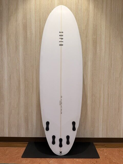 White surfboard with a central stringer, standing upright on a black display platform against a wood-paneled wall; brand name 'SUPFER' runs along the center.