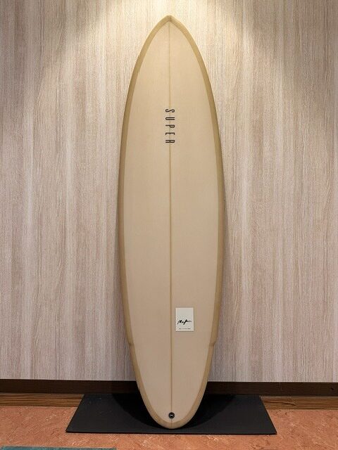 Cream-colored surfboard standing upright against a wood-paneled wall on a black display mat.